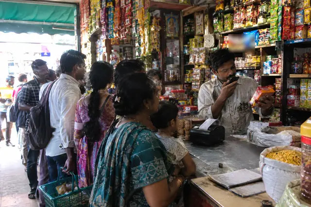 A shopkeeper at a busy grocery counter speaks into his phone while customers wait in line.