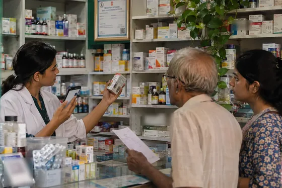 A pharmacist checks a medicine box with her phone while two customers wait at the counter.